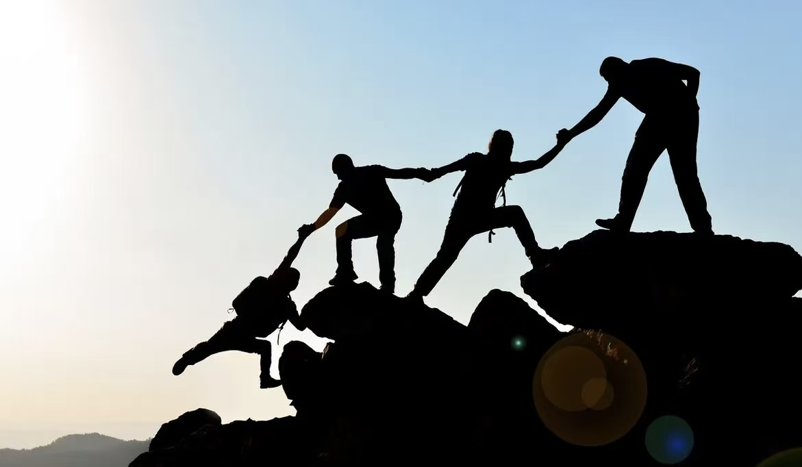 Silhouettes of climbers helping one another reach a rocky summit